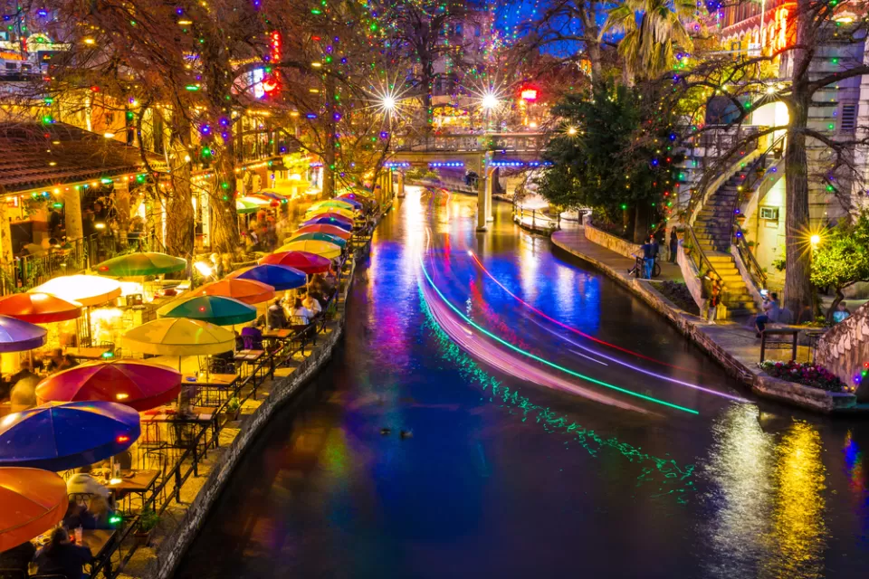View of San Antonio River Walk with colorful umbrellas at tables on waterfront and with boat lights blurred by long-exposure
